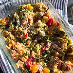 A glass dish filled with baked orzo pasta mixed with cherry tomatoes, green beans, red peppers, herbs, and grated cheese sits on a blue and white striped cloth.
