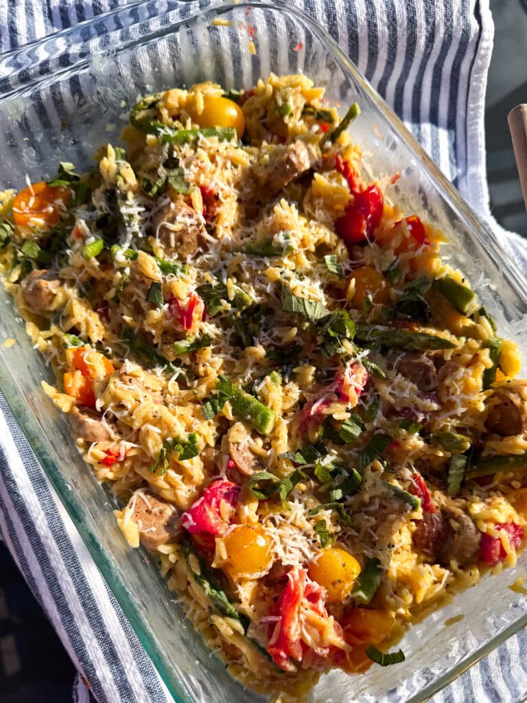 A glass dish filled with baked orzo pasta mixed with cherry tomatoes, green beans, red peppers, herbs, and grated cheese sits on a blue and white striped cloth.