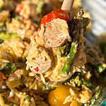 A close-up of a spoonful of creamy pasta with sliced sausage, asparagus, cherry tomatoes, and grated cheese, with the rest of the dish visible in the background.