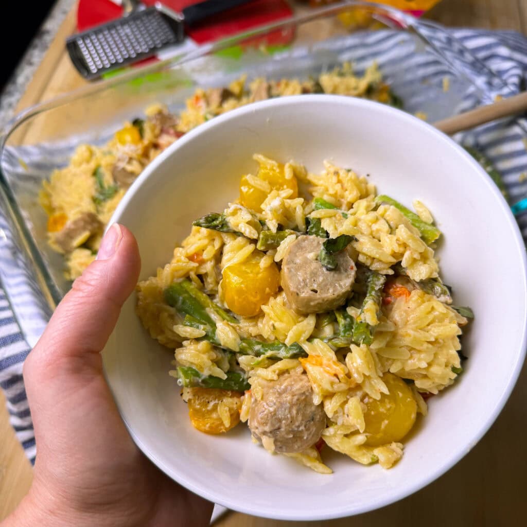 A hand holds a white bowl filled with creamy pasta, yellow cherry tomatoes, sausage slices, and asparagus; a larger baking dish of the same dish is in the background on a striped cloth.