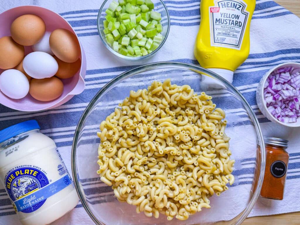 A bowl of cooked elbow macaroni surrounded by eggs, chopped celery, diced red onion, a jar of mayonnaise, a bottle of yellow mustard, and a container of paprika on a striped cloth.
