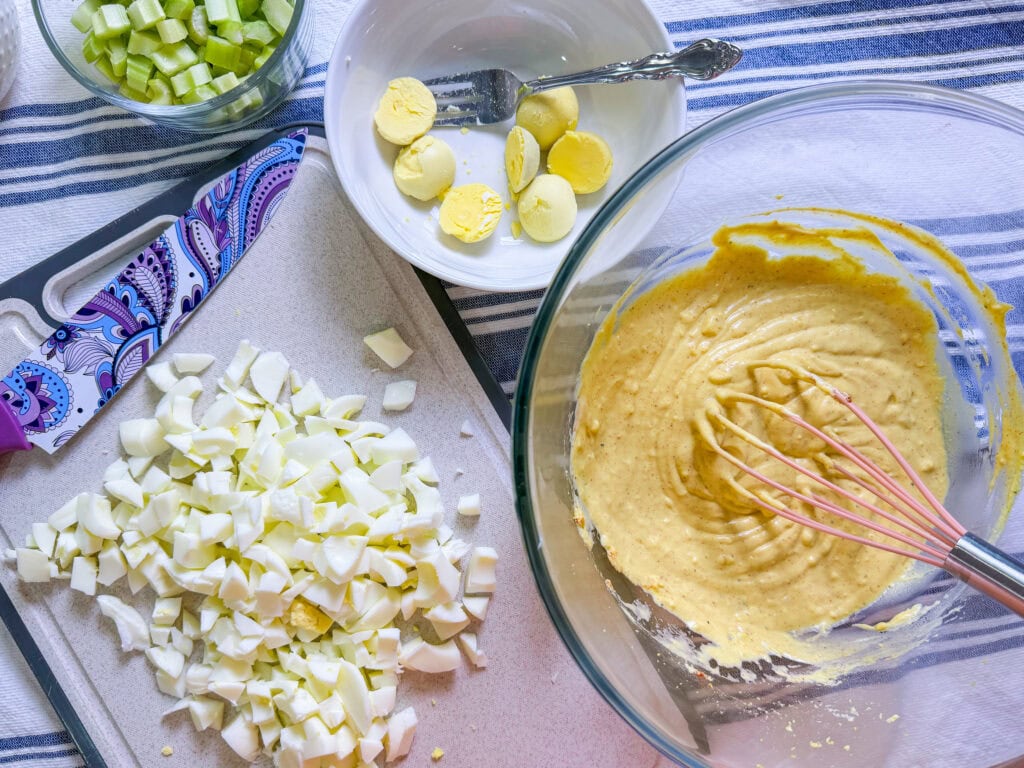 Chopped hard-boiled eggs on a cutting board, sliced celery in a bowl, hard-boiled egg halves on a plate, and a whisk in a bowl of yellow mixture, all on a striped cloth.