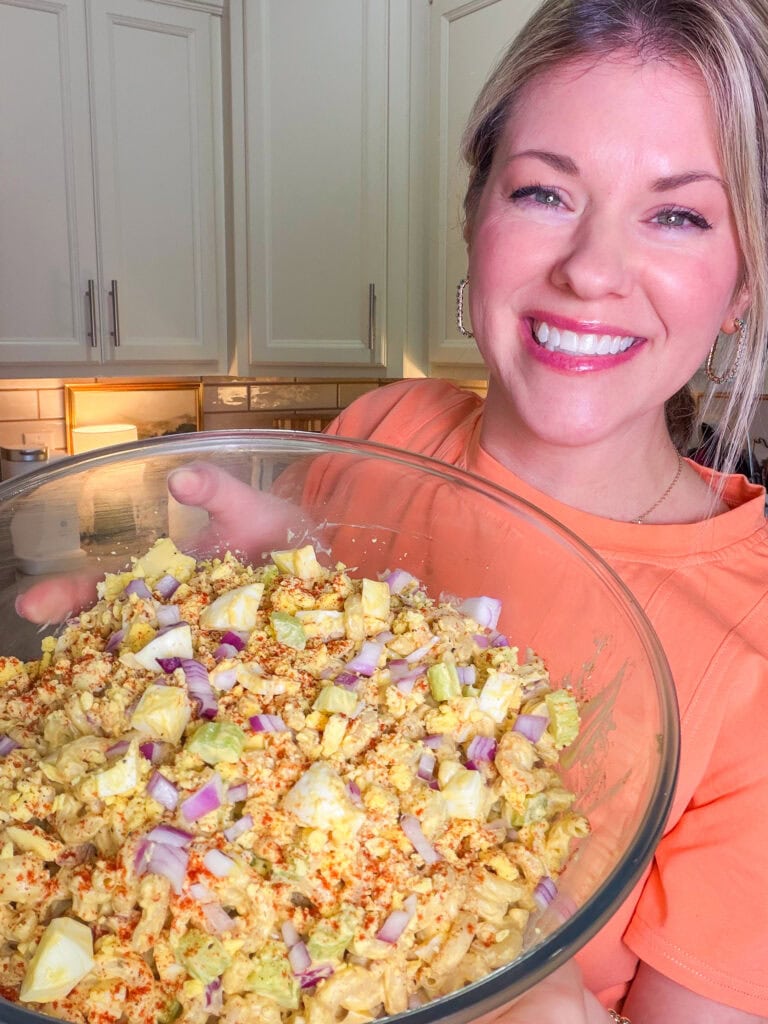A smiling woman in an orange shirt holds a large glass bowl filled with pasta salad topped with chopped red onions and sprinkled seasoning in a kitchen.
