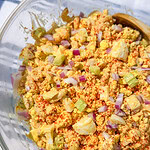 A close-up of a bowl of egg salad mixed with chopped red onions, celery, and sprinkled with paprika, in a clear glass bowl with a wooden spoon.