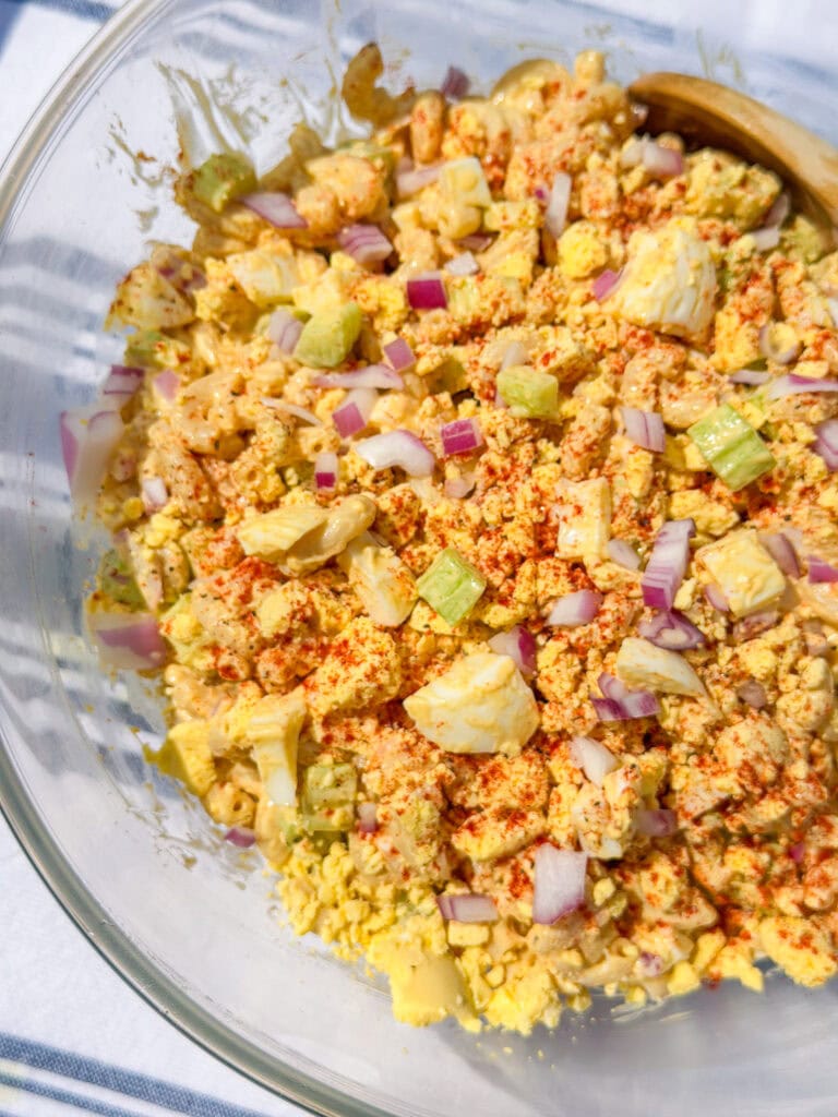A close-up of a bowl of egg salad mixed with chopped red onions, celery, and sprinkled with paprika, in a clear glass bowl with a wooden spoon.