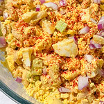 A close-up of macaroni salad with chopped hard-boiled eggs, diced celery, red onion, and a sprinkle of paprika mixed in a bowl.
