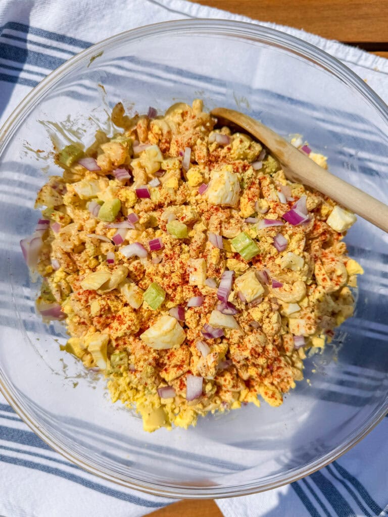 A clear glass bowl filled with egg salad mixed with chopped celery, red onion, and spices. A wooden spoon rests inside the bowl, which is placed on a white cloth with blue stripes.