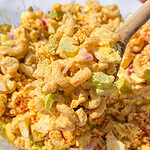 A close-up of a spoon scooping creamy macaroni salad with chopped celery, red onions, and hard-boiled eggs from a glass bowl, sprinkled with paprika.