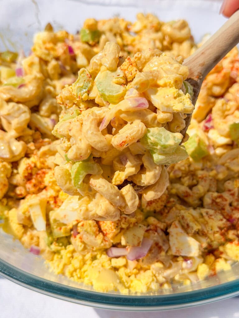 A close-up of a spoon scooping creamy macaroni salad with chopped celery, red onions, and hard-boiled eggs from a glass bowl, sprinkled with paprika.