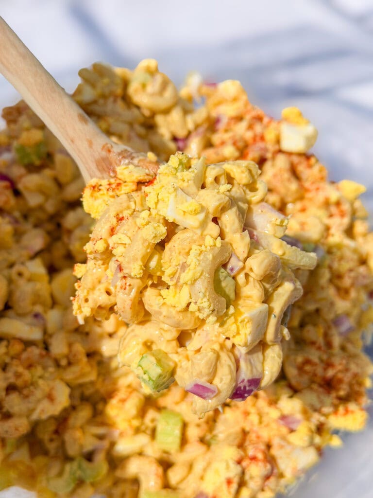 A close-up of a creamy macaroni salad with chopped eggs, celery, and red onion, being scooped with a wooden spoon. The salad is sprinkled with paprika.