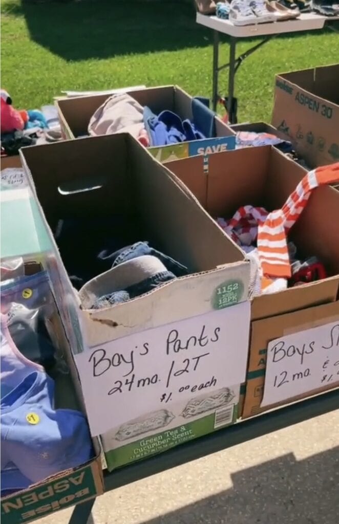 Cardboard boxes at an outdoor sale hold childrens clothes. A sign on one box reads Boys Pants 24 mo./2T $1.00 each, and another reads Boys Shirts 12 mo. Shoes and more boxes are visible nearby.