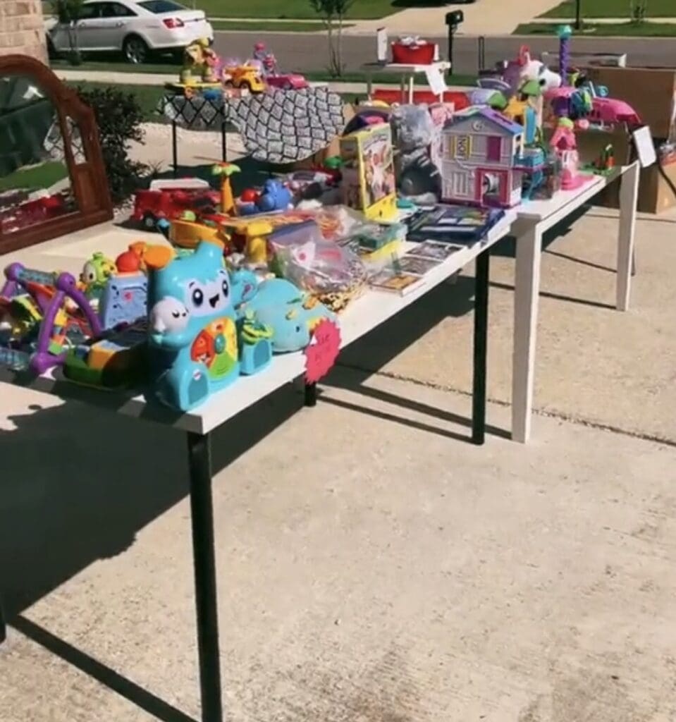 A yard sale setup on a driveway with tables displaying colorful childrenโs toys, games, and books; a car and more items are visible in the background on a sunny day.