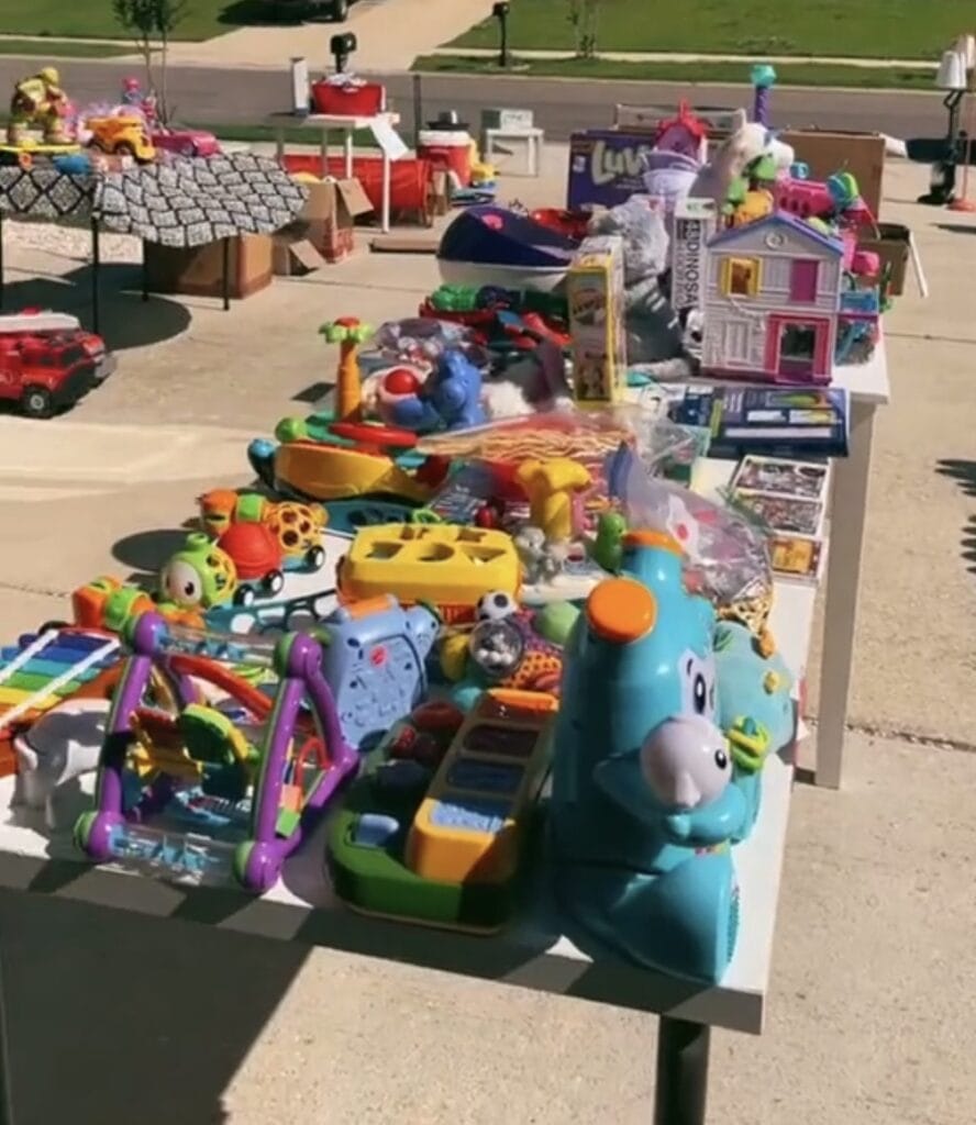 A table at a yard sale is filled with colorful childrens toys, including a blue hippo, playsets, xylophone, blocks, cars, and stuffed animals. More toys and items are visible on tables in the background.