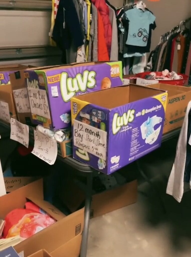 Several cardboard boxes labeled with signs, including diaper boxes, sit on a table in a garage sale setting. The boxes contain assorted baby clothes, and clothing racks with more clothes are visible in the background.