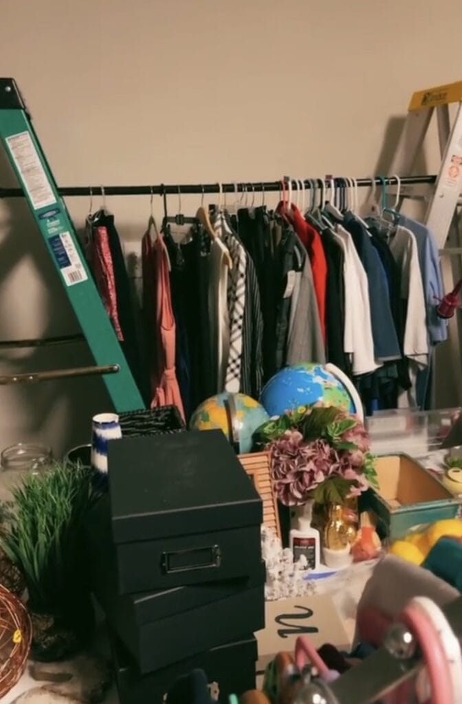 A cluttered room with a clothing rack holding various clothes, ladders on each side, stacked black boxes, two globes, artificial flowers, and miscellaneous household items scattered on a table in the foreground.