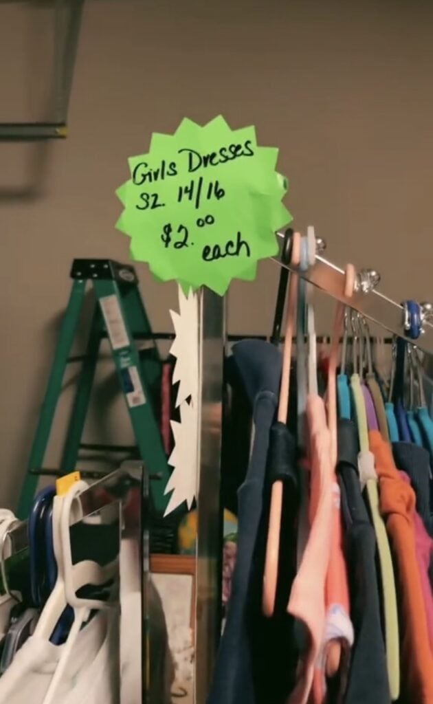 A bright green sign reading Girls Dresses Sz. 14/16 $2.00 each is attached to a rack of colorful dresses. A ladder is visible in the background.