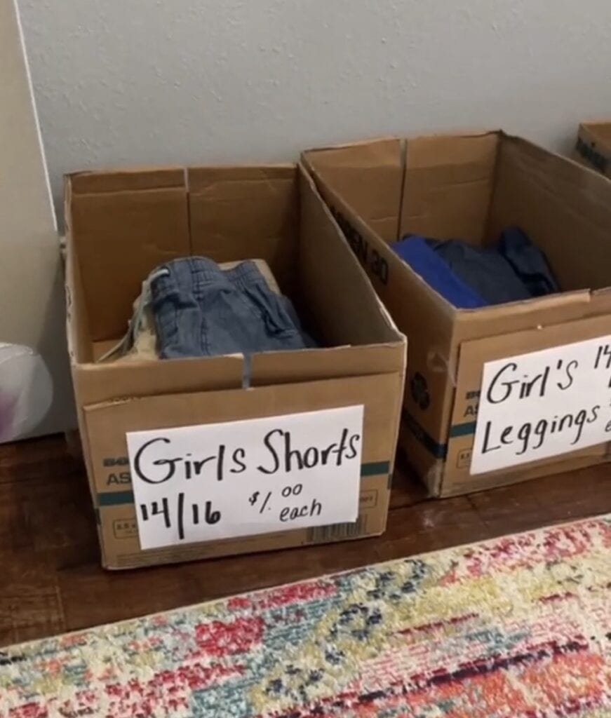 A cardboard box labeled Girls Shorts 14/16 $1.00 each sits on the floor, filled with folded shorts. Next to it is another box labeled Girls 14/16 Leggings, partially visible. A colorful rug is in the foreground.