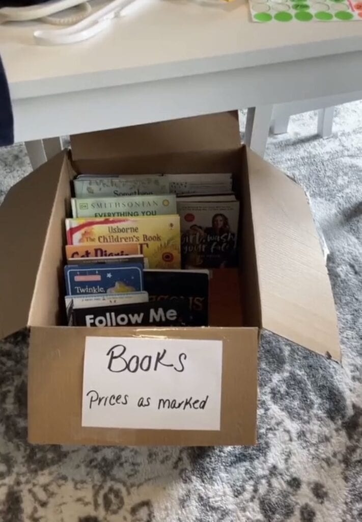 A cardboard box filled with childrens books and a sign on the front that reads Books Prices as marked, sitting on a gray patterned carpet near a white table.