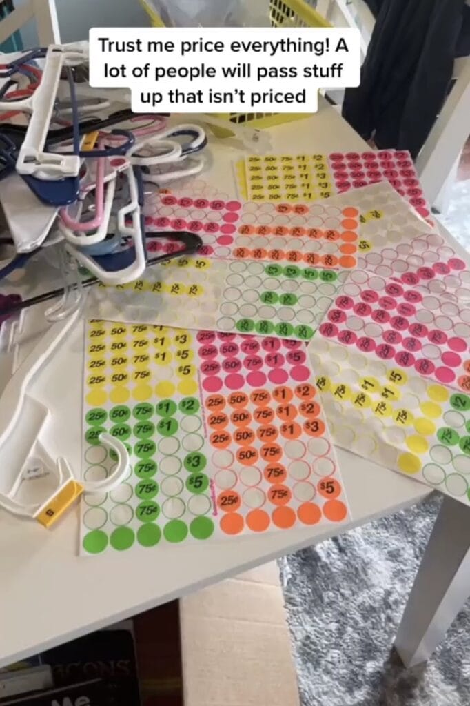 Sheets of colorful price stickers labeled with various amounts are spread out on a table. Nearby are plastic hangers and a pricing tag gun. A caption above reads, Trust me price everything! A lot of people will pass stuff up that isnโt priced.