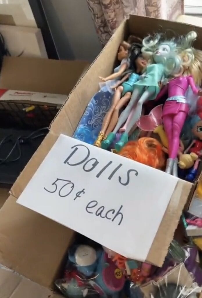 A cardboard box filled with various dolls, including some with colorful outfits and hair. A handwritten sign on the box reads Dolls 50ยข each. Other items are partially visible in the background.