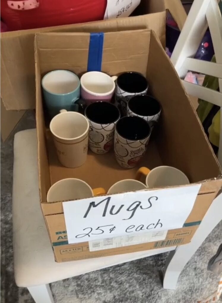 A cardboard box labeled Mugs 25ยข each sits on a white chair, containing various mugs of different colors and patterns. The box is open and placed indoors on a carpeted floor.