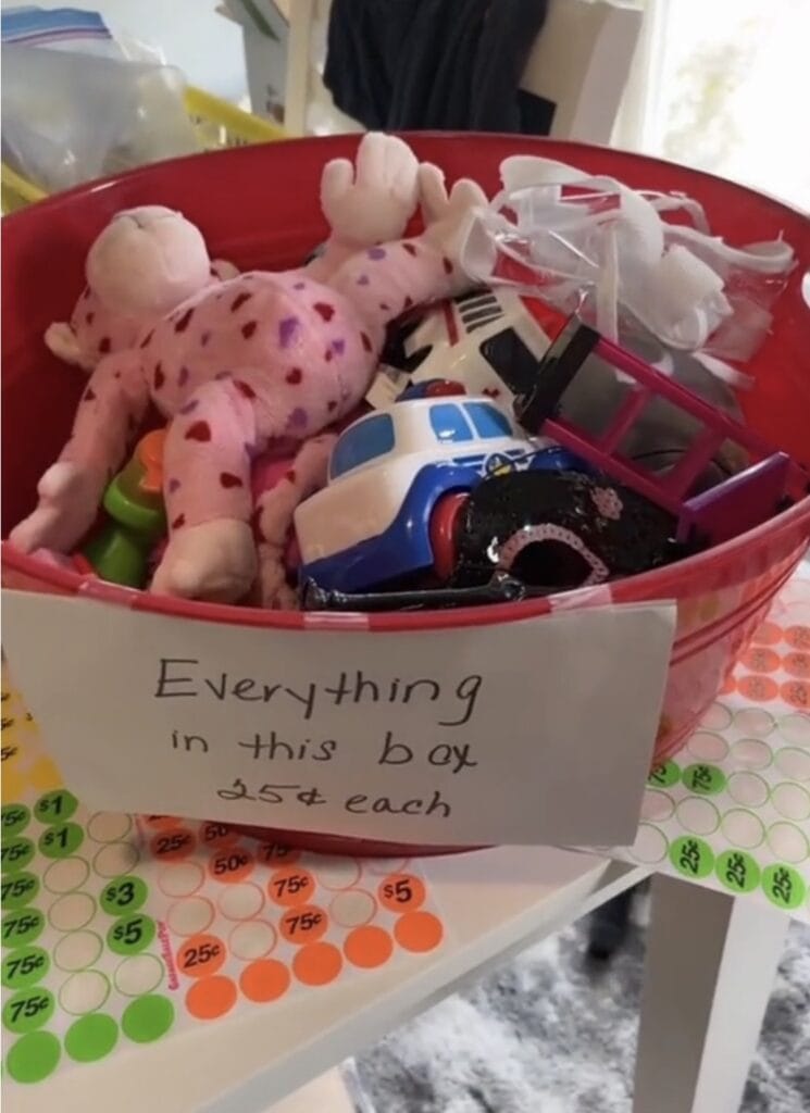 A red container filled with small toys, including a pink stuffed animal, cars, and other items. A handwritten sign on the box reads, Everything in this box 25ยข each. The box sits on a table with colorful price stickers.
