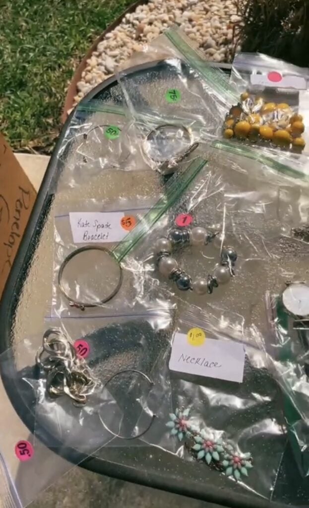 Assorted jewelry pieces, including bracelets, a necklace, and earrings, individually bagged with handwritten price tags and colorful stickers, displayed on a table outdoors at what appears to be a yard sale.