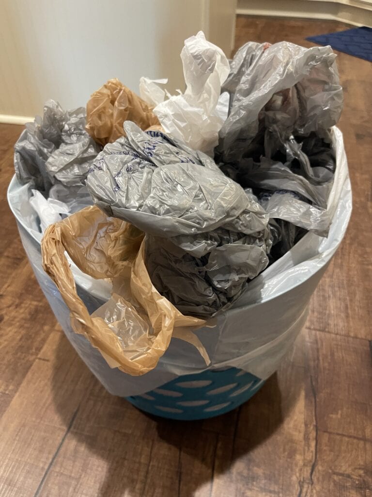 A blue plastic basket is filled to the brim with various crumpled plastic bags in gray, white, and brown colors. The basket sits on a wooden floor near a hallway entrance.