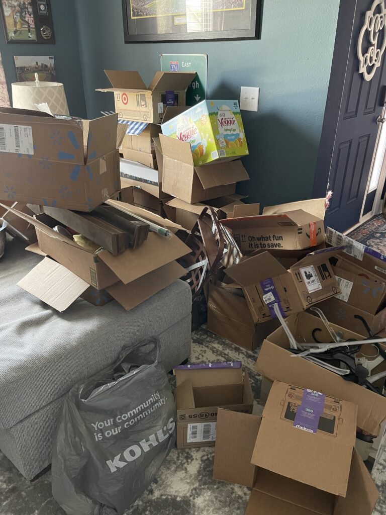 A large pile of empty cardboard boxes and packaging materials stacked messily next to a gray couch, with a Kohlโs shopping bag on the floor, in a cluttered living room near a dark blue door.