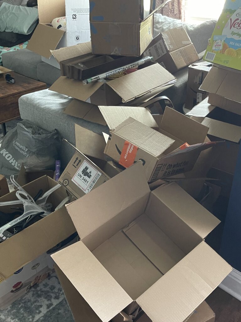 A large pile of open and flattened cardboard boxes, some containing various household items, sits on a rug in a living room near a gray sofa and a wooden table.