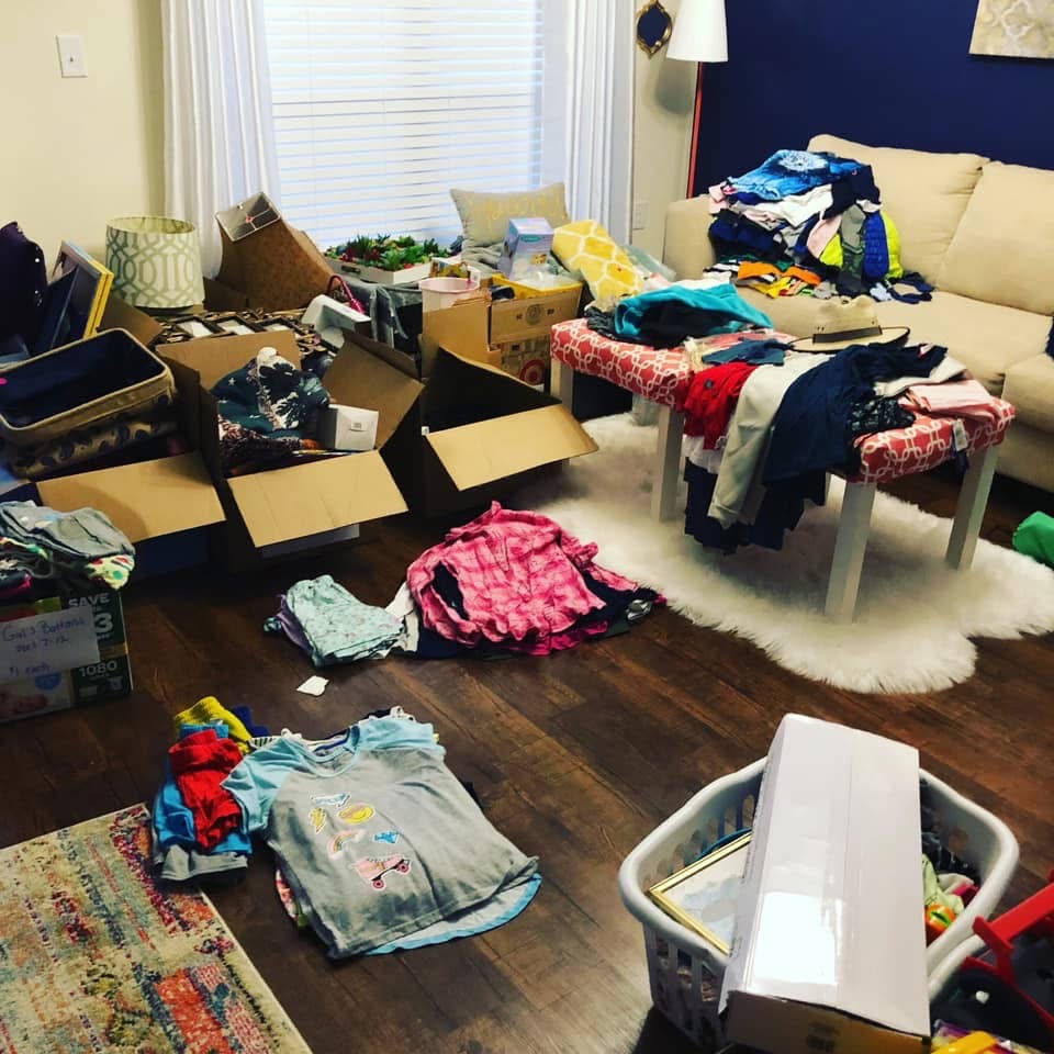 A living room with piles of clothes, cardboard boxes, and laundry baskets spread across the floor and coffee table. A sofa and window are in the background, and the area looks cluttered and busy with sorting or organizing.