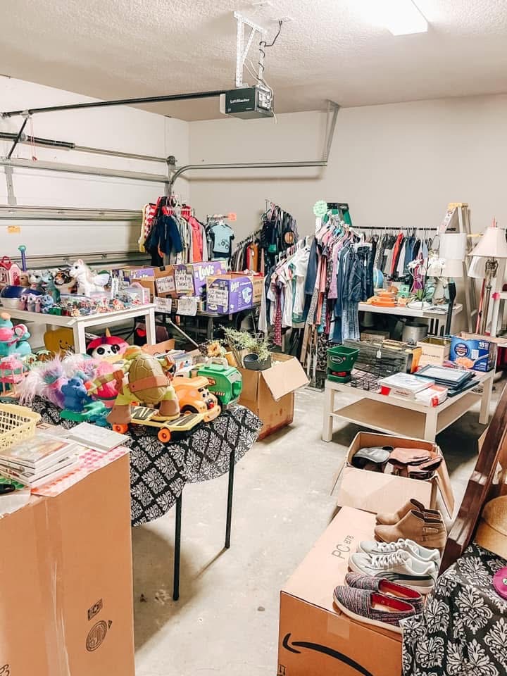 A garage filled with tables and racks displaying clothes, toys, shoes, books, and household items for a garage sale. Cardboard boxes and bins are scattered around the organized space.