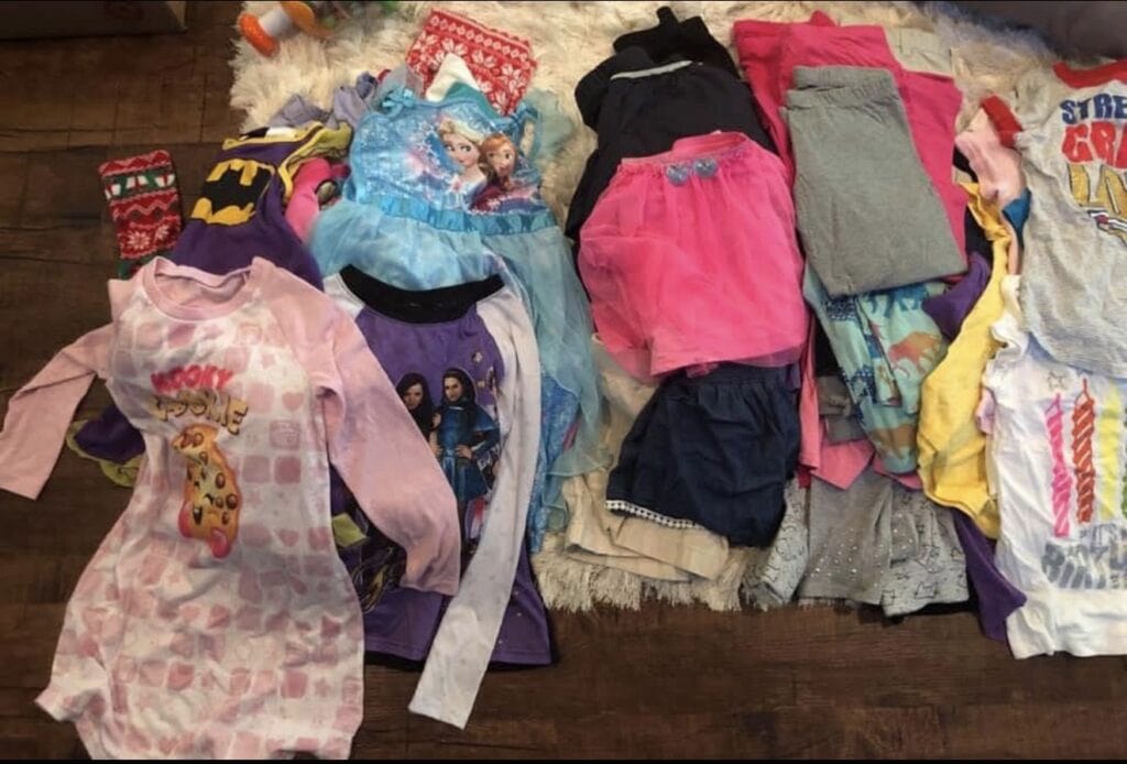 A variety of childrens clothes are laid out on a white rug and wooden floor, including pajamas with cartoon characters, dresses, T-shirts, skirts, and shorts in different colors and patterns.