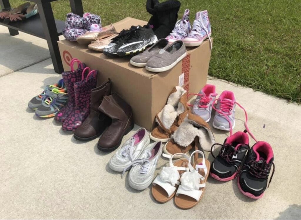 A variety of childrenโs shoes, including sneakers, sandals, boots, and slippers, are neatly arranged on and around cardboard boxes outdoors on a sunny day next to a green lawn.