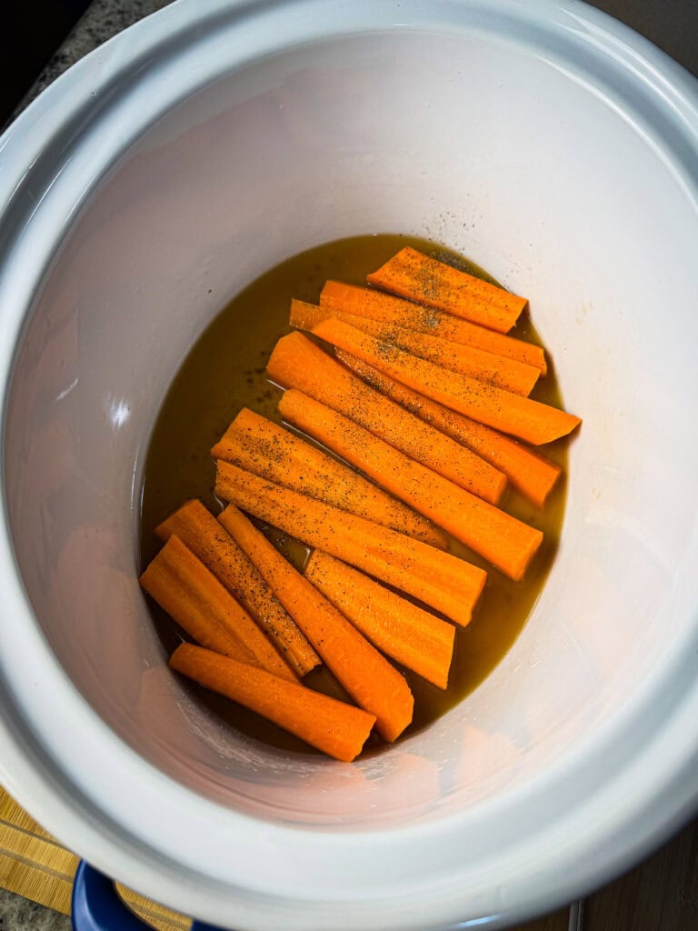 Carrot sticks seasoned with black pepper are placed in a slow cooker with a small amount of liquid at the bottom, ready for cooking.