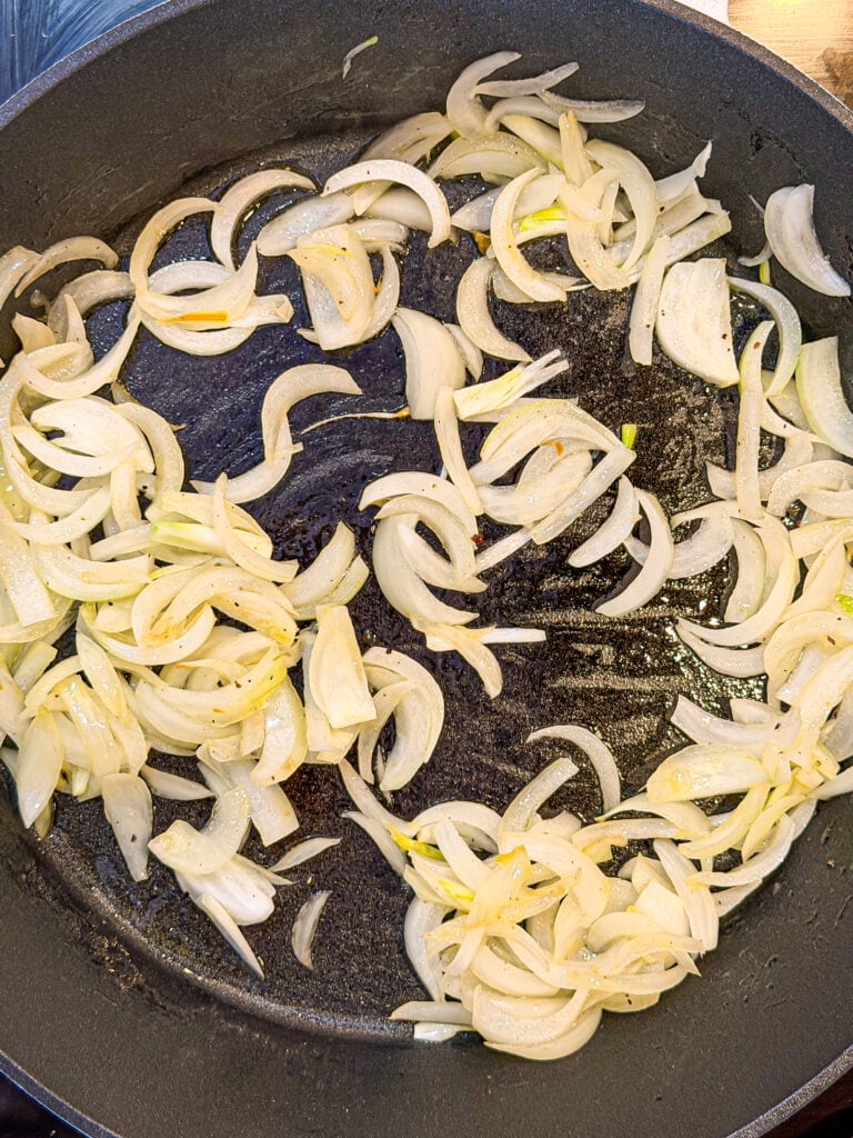 Sliced onions cooking in a black skillet, starting to soften and turn translucent with a light sheen of oil visible on the surface.