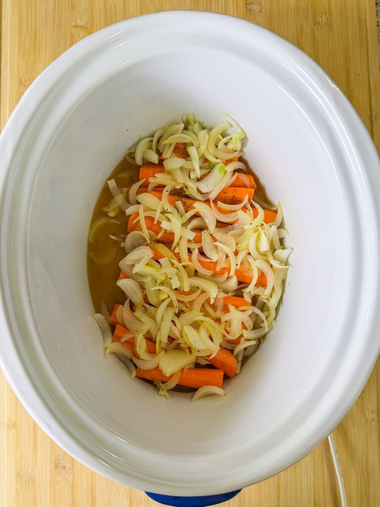 Sliced onions and carrots are placed in a white slow cooker with broth, ready for cooking, on a light wooden surface.