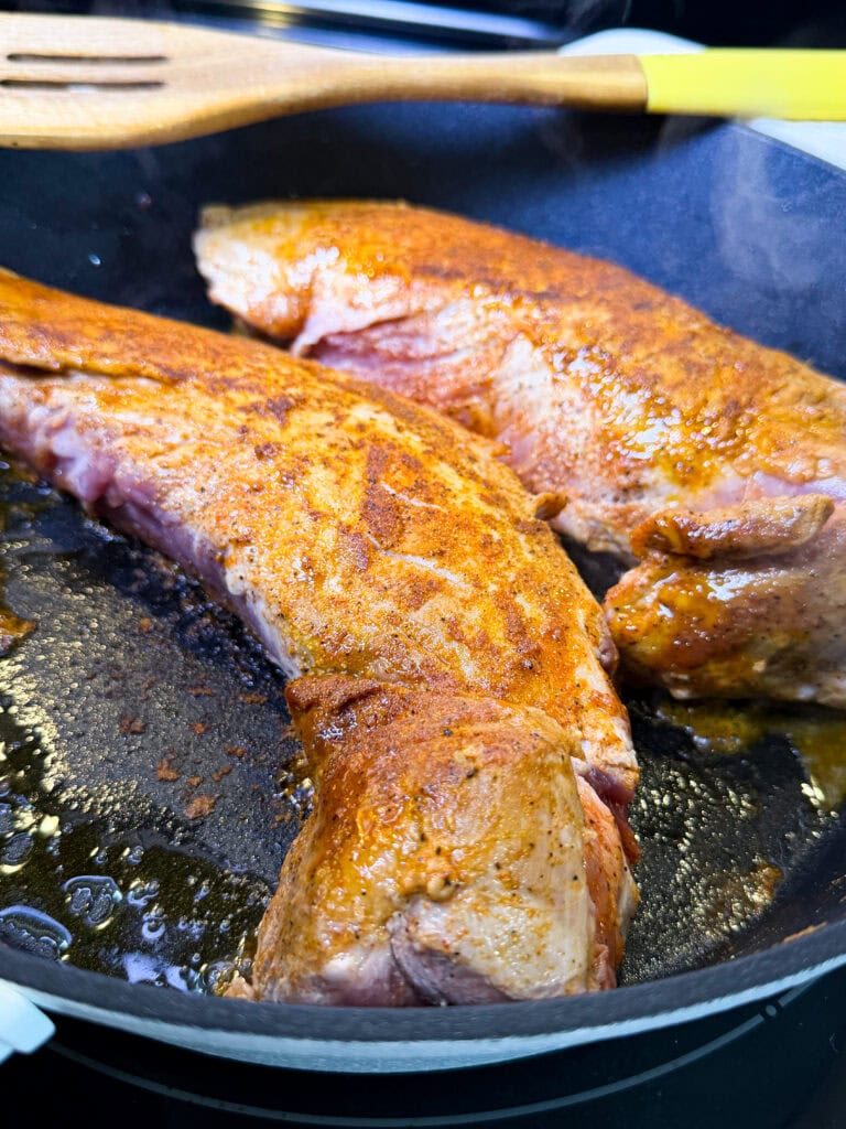 Two pieces of seasoned pork tenderloin are being seared in a black skillet, with a wooden spatula resting on the edge of the pan. The meat is lightly browned and sizzling in oil.