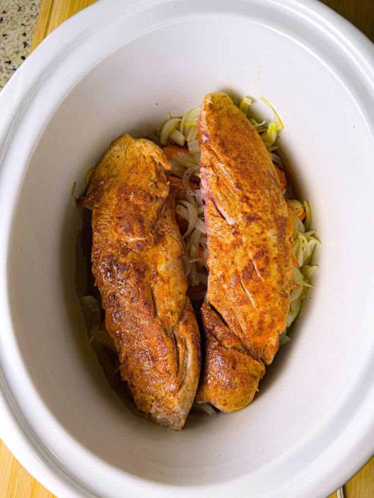 Two seasoned pork tenderloins sit on top of sliced onions and vegetables inside a white slow cooker, ready to be cooked.