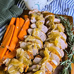 Sliced pork tenderloin topped with onion gravy on a wooden board, served with carrot sticks. Fresh rosemary sprigs and a cup of extra gravy are beside the meat. A striped cloth napkin is in the background.