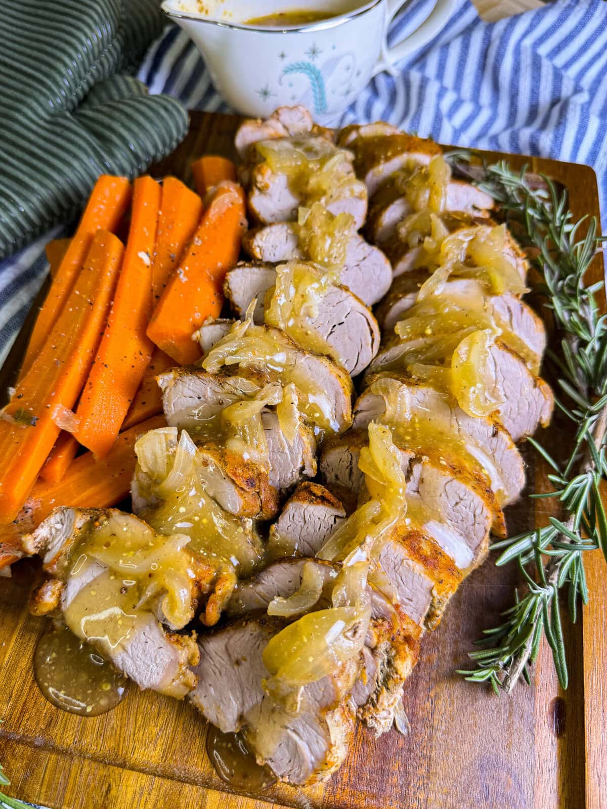 Sliced pork tenderloin topped with onion gravy on a wooden board, served with carrot sticks. Fresh rosemary sprigs and a cup of extra gravy are beside the meat. A striped cloth napkin is in the background.