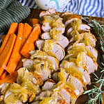 Sliced pork tenderloin topped with onion gravy is arranged on a wooden board, served with cooked carrot sticks, fresh rosemary sprigs, and a side of stacked green plates.