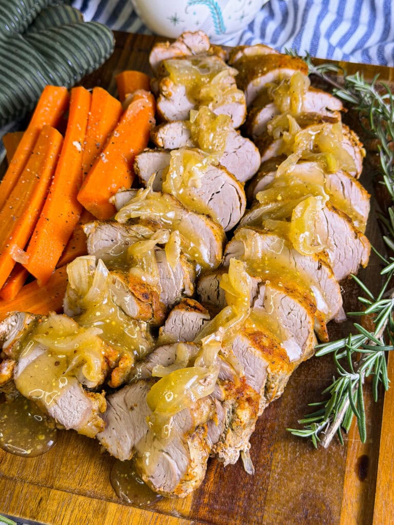 Sliced pork tenderloin topped with onion gravy is arranged on a wooden board, served with cooked carrot sticks, fresh rosemary sprigs, and a side of stacked green plates.