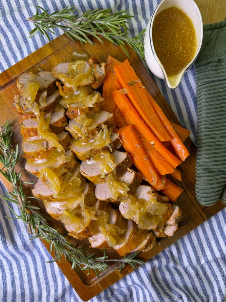 A wooden board with sliced pork tenderloin topped with caramelized onions, roasted carrots, fresh rosemary sprigs, and a small pitcher of gravy, all placed on a blue and white striped cloth.