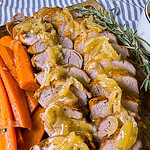 Sliced pork tenderloin topped with caramelized onions and gravy, served on a wooden board with cooked carrot sticks and garnished with fresh rosemary sprigs.