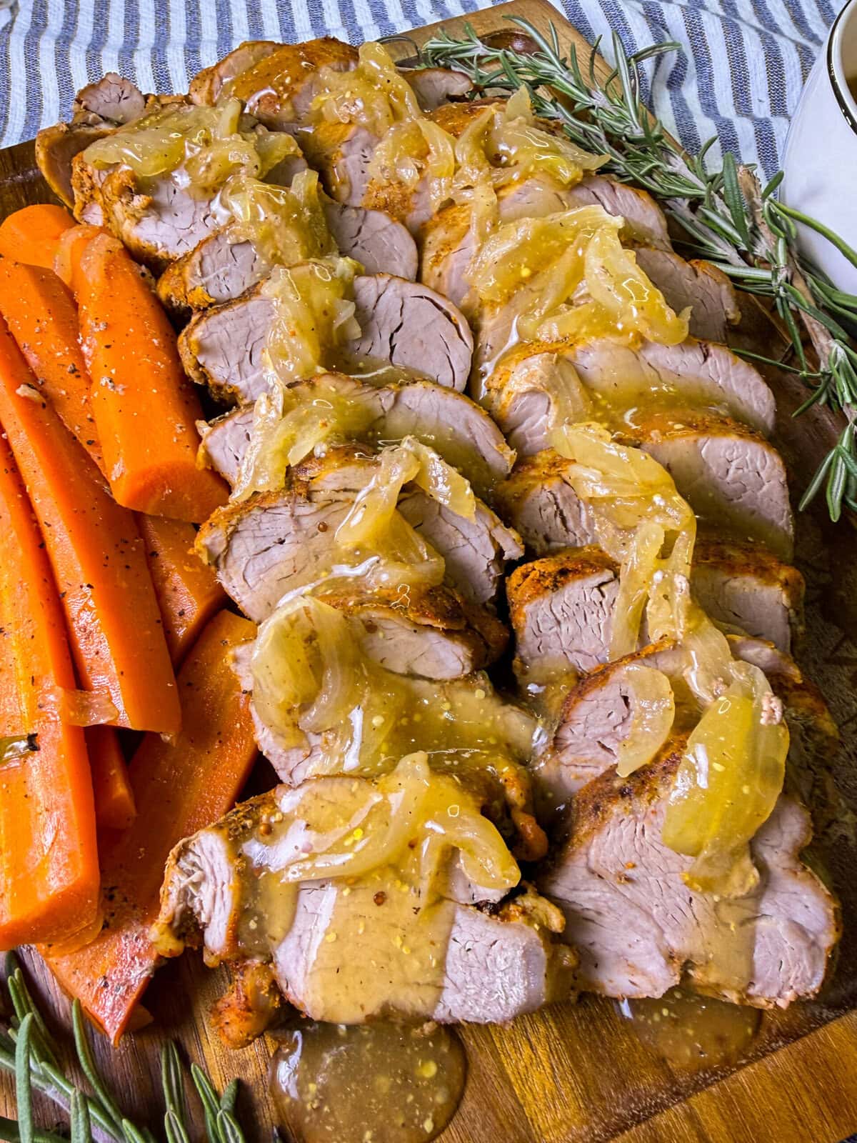 Sliced pork tenderloin topped with caramelized onions and gravy, served on a wooden board with cooked carrot sticks and garnished with fresh rosemary sprigs.