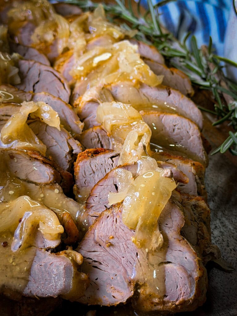 Sliced roasted pork tenderloin topped with caramelized onions and a glossy sauce, garnished with a sprig of fresh rosemary on the side.