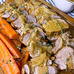 Sliced roast pork tenderloin topped with onion gravy, garnished with fresh herbs, served alongside cooked carrot sticks on a wooden board; a gravy boat sits in the background.