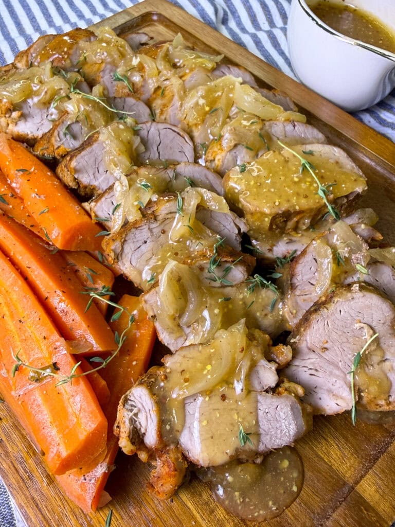 Sliced roast pork tenderloin topped with onion gravy, garnished with fresh herbs, served alongside cooked carrot sticks on a wooden board; a gravy boat sits in the background.