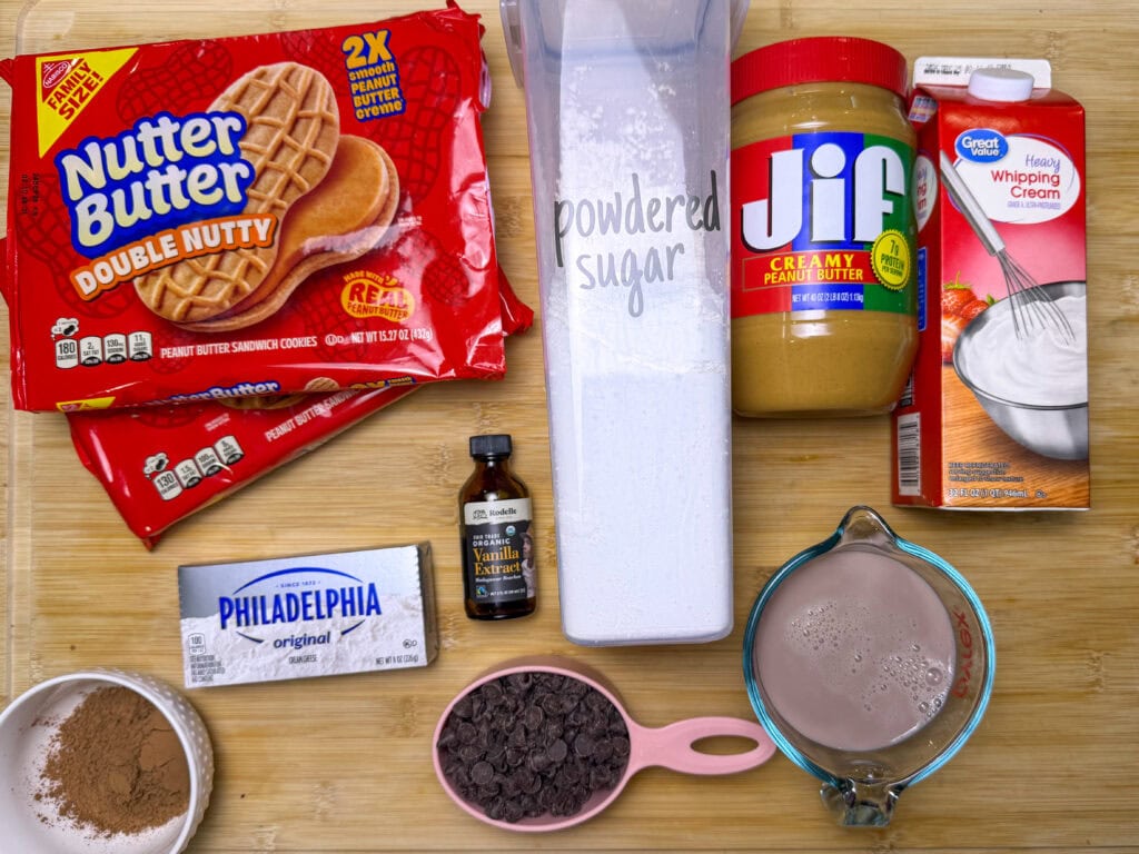 Ingredients on a wooden surface include Nutter Butter cookies, powdered sugar, Jif peanut butter, heavy whipping cream, Philadelphia cream cheese, cocoa powder, vanilla extract, chocolate chips, and chocolate milk.